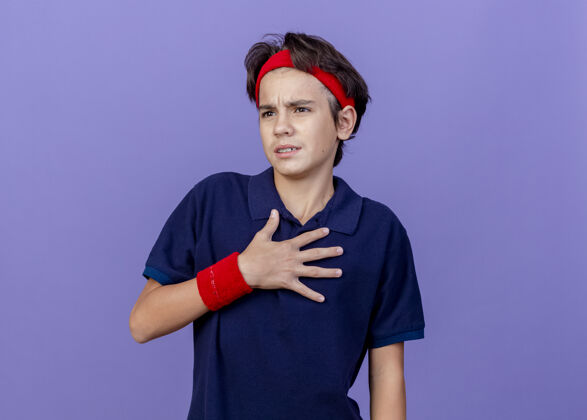 牙齿皱眉的年轻帅气的运动男孩戴着头带和护腕 戴着牙套 看着侧面 手放在胸前 隔离在紫色的墙上背带人人
