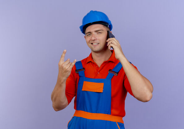 年轻身着制服 头戴安全帽的年轻男性建筑工人在电话里高兴地说着话 并在紫色地板上展示山羊的姿势电话手势山羊