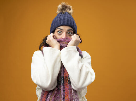 疾病印象深刻的年轻生病的女人穿着长袍冬季帽子和围巾看着前面的遮嘴围巾隔离在橙色的墙上年轻人感情冬天
