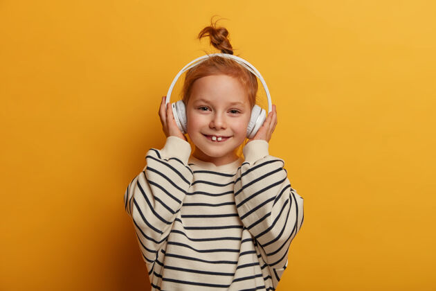 小小女孩梅洛曼戴着新耳机享受着美妙的声音 听着最喜欢的歌 听着咕噜音乐 露出乳牙 穿着条纹套头衫 孤零零地站在黄色的墙上 有着无忧无虑的心情发髻声音音乐