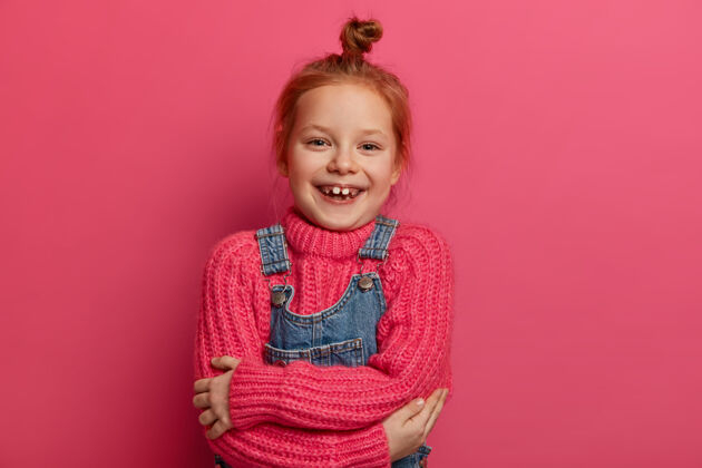 女学生开朗的小姜姑娘抱着自己 感觉很舒服 有一件新的羊毛粉色毛衣 暖和柔软的衣服 笑容爽朗 露出缺牙 有一头姜黄色的头发 孤立在粉色的墙上结套头衫快乐