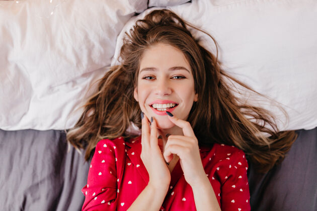 卧室优雅的白人女孩躺在床上表达积极的情绪快乐的棕色头发女人微笑的室内照片快乐肖像寒冷