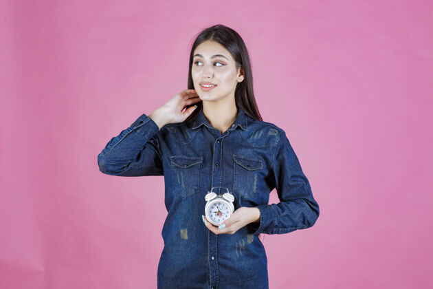 人一个穿着牛仔衬衫的年轻女人拿着一个闹钟聪明休闲姿势