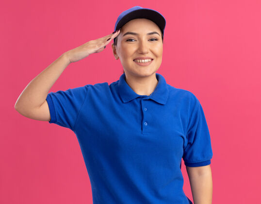 站立身着蓝色制服 头戴帽子的快乐的年轻女送货员站在粉红色的墙上 微笑着自信地向前面敬礼微笑自信制服