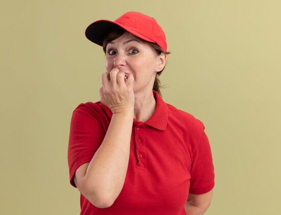 紧张身穿红色制服 头戴鸭舌帽的中年女送货员站在绿色墙壁上 看着前面紧张紧张 咬牙切齿的钉子分娩制服中年