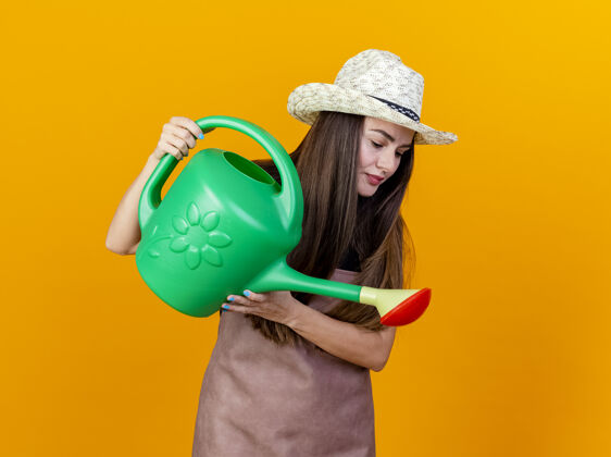 帽子看着美丽的园丁女孩穿着制服 戴着园艺帽 用浇水器浇水 可以隔离在橙色的背景上园艺女孩羽绒