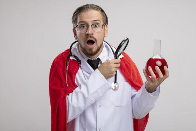填补年轻的超级英雄穿着医用长袍 戴着听诊器和眼镜 手里拿着装满红色液体的chesmistry玻璃瓶 放大镜隔离在白色背景上医疗听诊器惊喜