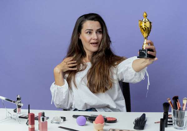 紫色惊讶的美丽女孩坐在桌子旁 拿着化妆工具 看着紫色墙上孤立的优胜者杯拿着坐着化妆