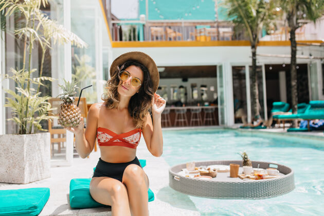 热带戴着太阳镜的迷人卷发女人在度假胜地享用鸡尾酒阳光明媚的日子里 美丽的棕褐色女人拿着菠萝坐在游泳池旁的户外照片太阳笑模特