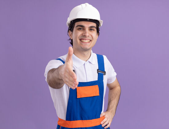 建设者身穿施工制服 头戴安全帽的年轻建筑工人站在紫色的墙上 面带微笑 友好地举手致意头盔年轻人男人