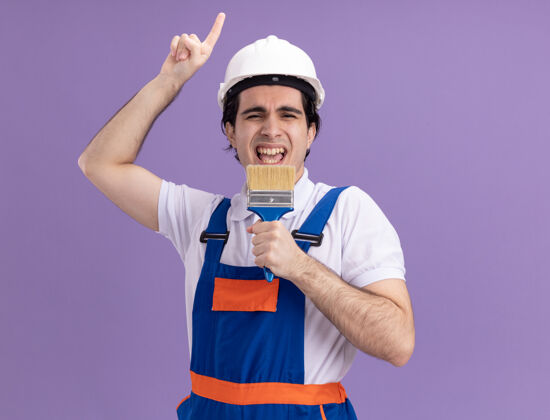 抱着年轻的建筑工人身穿建筑制服 戴着安全帽 手里拿着油漆刷 站在紫色的墙上 快乐地唱着歌建筑建设者快乐