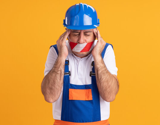 手疼痛的白种人成年建筑工人穿着制服 嘴上缠着胶带 手放在橘黄色的太阳穴上疼痛制服建筑工人