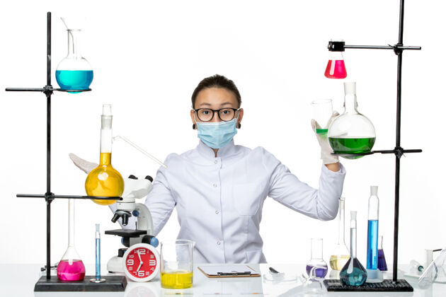 专业前视图穿着医疗服的女化学家戴着口罩拿着绿色溶液在白色地板上飞溅实验室病毒化学covid-病毒医生面具