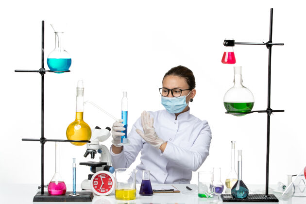 喷溅前视图穿着医疗服的女化学家带着面罩拿着浅白色背景上蓝色溶液的烧瓶溅起病毒化学实验室的病毒正面面罩套装
