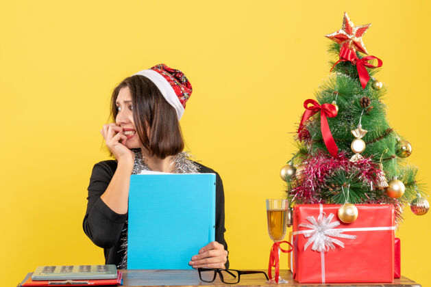 圣诞老人一位穿着西装 戴着圣诞老人帽和新年装饰品 手里拿着文件的迷人女士在黄色的办公室里与世隔绝黄色快乐成人