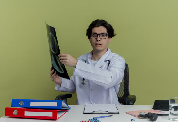 人年轻的男医生穿着医用长袍 戴着听诊器 戴着眼镜 坐在办公桌旁 手里拿着医疗工具 手里拿着x光片 看上去很孤立听诊器镜头年轻人
