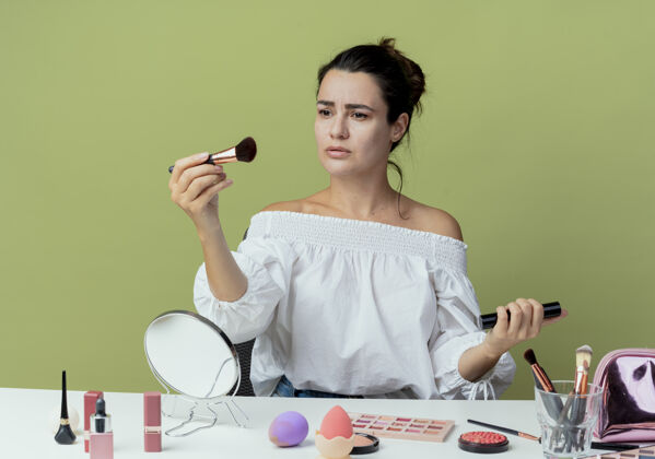 化妆美丽的心烦意乱的女孩坐在桌子旁 拿着化妆工具 把刷子和睫毛膏隔离在绿色的墙上刷子拿着女人