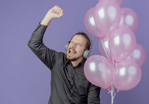 升起戴着耳机的快乐帅哥手持氦气球 在紫色的墙上举起拳头生日氦气拳头