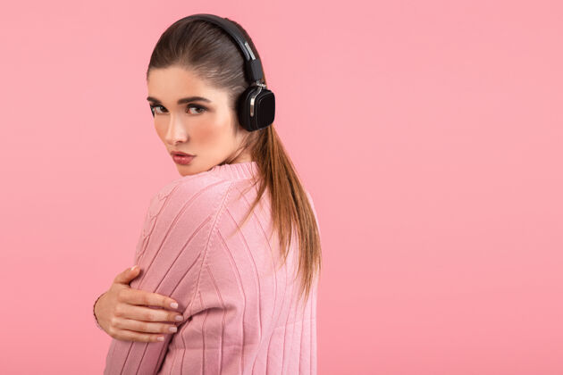 年轻年轻迷人的女人戴着无线耳机听音乐穿着粉色毛衣微笑着快乐积极的心情摆在粉色背景上孤立乐趣漂亮时髦