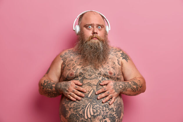 胖乎乎惊讶的家伙一直把手放在肚子上 在肚子和胳膊上纹身 赤裸着身体站着 超重 用耳机听动听的音频 隔离在粉红色的墙上奇迹腹部播放列表