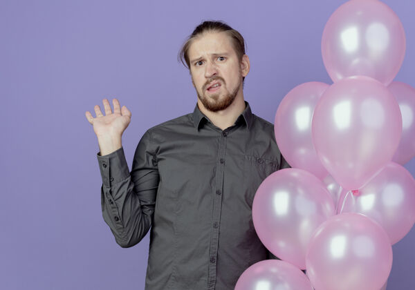 氦恼怒的帅哥拿着氦气球 举起手孤立地站在紫色的墙上起来拿着手