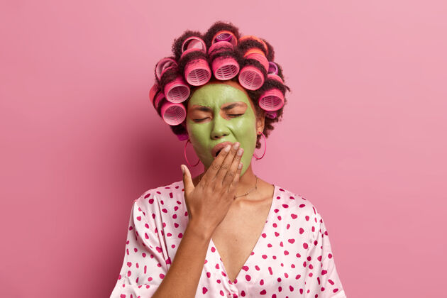 年轻家庭主妇的画像一早醒来 有美容程序捂嘴打哈欠 穿丝质长袍 头上用卷发器孤立过粉色发型 健康理念室内水疗美容