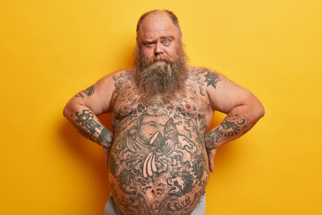 体重大胡子胖子有浓密的胡须 纹身的身体和大肚子 从眉毛下面看 手放在腰上 隔离在黄色的墙壁上肥胖 抽脂 减肥概念男人身材腹部