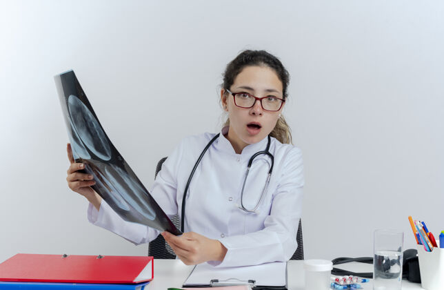 长袍年轻的女医生穿着医用长袍 戴着听诊器 戴着眼镜 坐在办公桌旁 手里拿着医疗工具 手里拿着x光片 看上去很孤立X光女士穿着