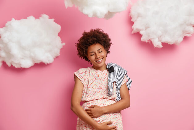妈妈非常快乐微笑的非洲裔美国孕妇轻轻摸肚子 摸宝宝 给新生儿买连身衣 享受孕产妇的时刻 与珍贵的未来胎儿玩耍肚子快乐身体