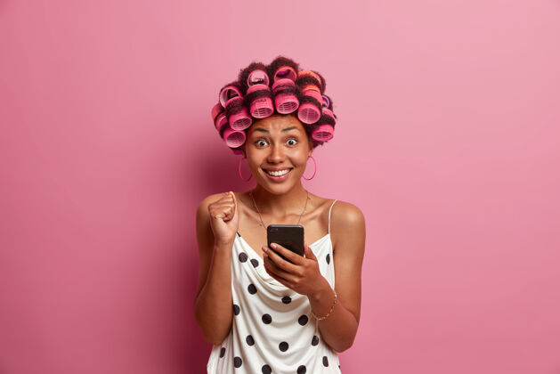 在线快乐的黑皮肤女人的肖像在智能手机上阅读精彩新闻 举起紧握的拳头和微笑 应用卷发器做发型家庭主妇在家使用社交网络女人电话滚轴