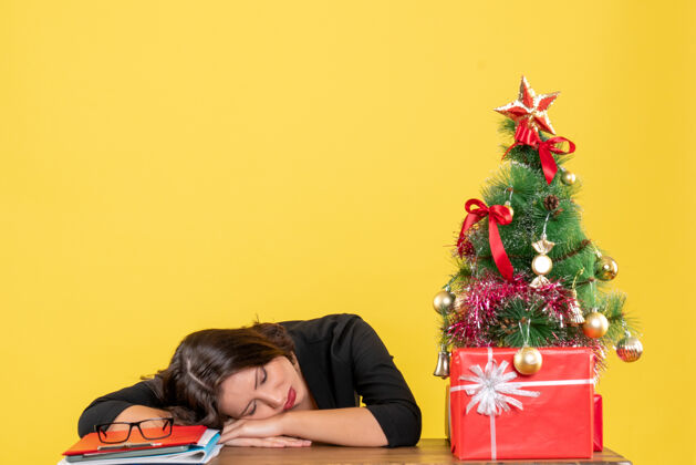 礼物在黄色的办公室里 一个年轻的女人坐在装饰过的圣诞树旁的桌子旁睡觉人节日礼物