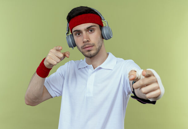 手臂戴着头戴式耳机和智能手机臂带的年轻运动型男子站在绿色背景下 手指指向相机 看起来很自信运动耳机立场