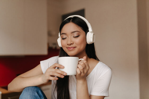 应用程序迷人的亚洲女人戴着耳机吸入茶香 捧着杯子微笑着设备咖啡舒适