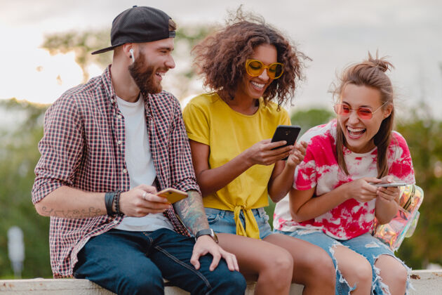 享受快乐的年轻朋友们坐在公园里用智能手机微笑 男人和女人玩得开心教育眼镜音乐