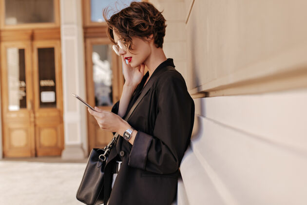 阳光穿着黑色夹克的短发酷女人在户外拿着平板电脑的资料照片戴着眼镜的漂亮女人拿着手提包在街上摆姿势年轻单身姿势