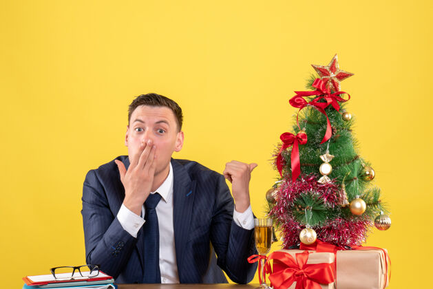 礼物前视图惊讶的男人手指圣诞树坐在桌子旁圣诞树和礼物上黄色的背景人男主管
