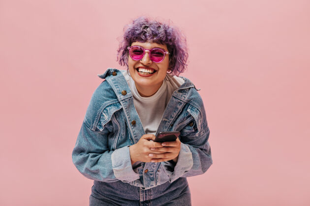 太阳镜明亮的卷发女人 淡紫色头发 戴着时髦的粉色眼镜 牛仔夹克和牛仔裤 手里拿着她的智能手机粉色女人短发