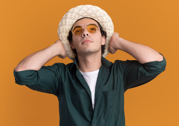 立场一个穿着绿色衬衫 戴着夏天帽子 戴着眼镜的年轻人站在橘色的墙上 脸上露出自信的表情表情穿着眼镜