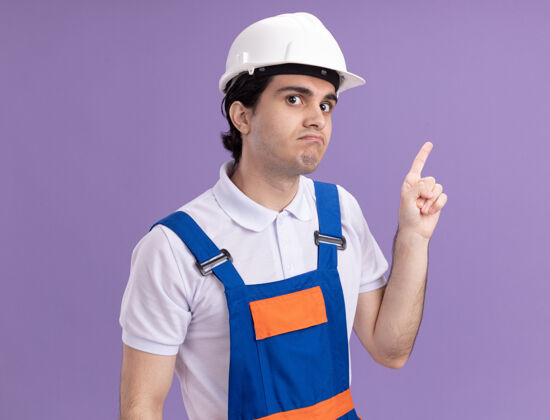 目录年轻的建筑工人穿着建筑制服 戴着安全帽 看上去很困惑 用食指指着站在紫色墙上的一边混乱建设者制服
