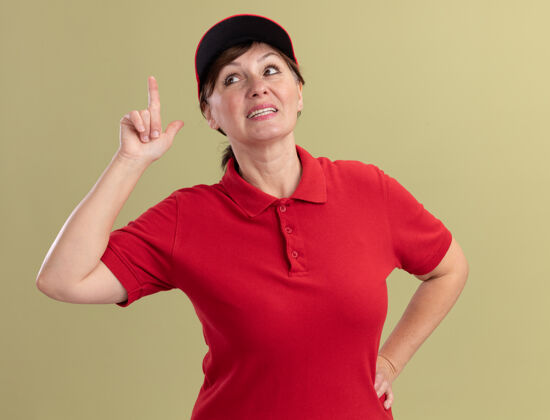 自信身着红色制服 头戴鸭舌帽的中年女送货员站在绿色的墙边 微笑着自信地展示着有新创意的食指微笑帽子手指