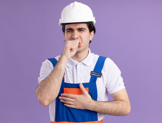 建筑工人身穿施工制服 头戴安全帽的年轻建筑工人站在紫色的墙上咳嗽 看上去很不舒服建筑安全头盔