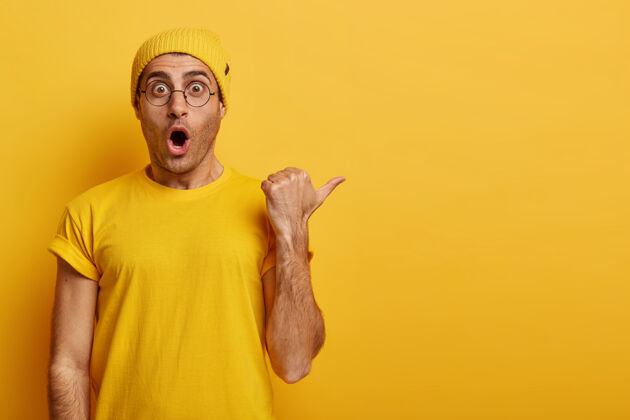 印象哇 巨大的折扣机会表情惊恐的情绪化男人展示了意想不到的机会 大拇指指向空白处 穿着黄色服装广告可怕眼镜旁白