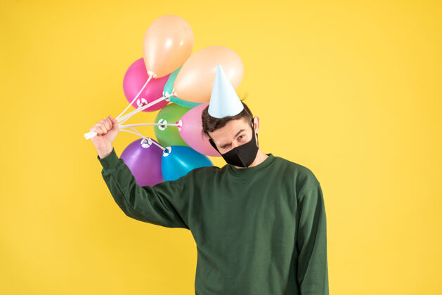 男人正面图：戴派对帽的年轻人手持五颜六色的气球站在黄色背景上卡通站立剪影