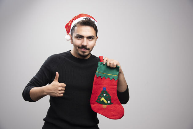 情感一个戴着圣诞老人帽子的年轻人竖起大拇指 手里拿着圣诞袜子男孩圣诞老人袜子