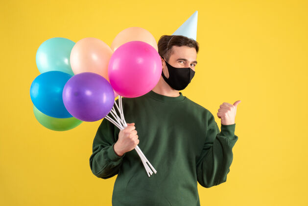 前面正面图：戴着派对帽的年轻人拿着气球站在黄色背景上聚会背景举行