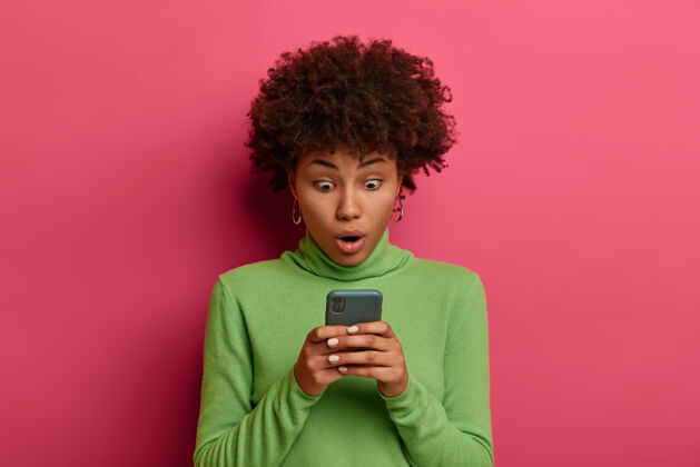 聊天科技和情感概念惊呆了的黑皮肤女人使用现代手机 盯着显示器 惊讶于好的小玩意 在线聊天高领毛衣卷发信息