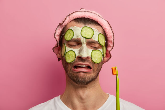 皮肤护肤治疗理念悲哀不满的男人伤心地看着牙刷 留着胡茬 用黄瓜敷面膜治疗干净阳刚