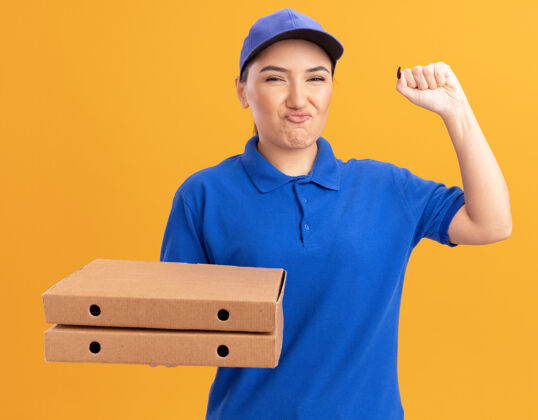 女人身着蓝色制服 头戴鸭舌帽 拿着比萨饼盒的年轻送货员高兴地看着前面 微笑着高举拳头 就像一个站在橙色墙壁上的胜利者送货请喜欢