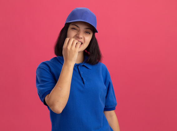 年轻穿着蓝色制服 戴着帽子的年轻送货员紧张地站在粉红色的墙上咬指甲咬人站立制服