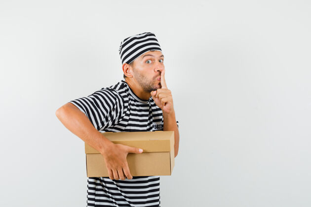 帽子一个年轻人拿着一个纸板箱 戴着条纹t恤帽子 表现出沉默的姿态 看上去很小心成功盒子现代